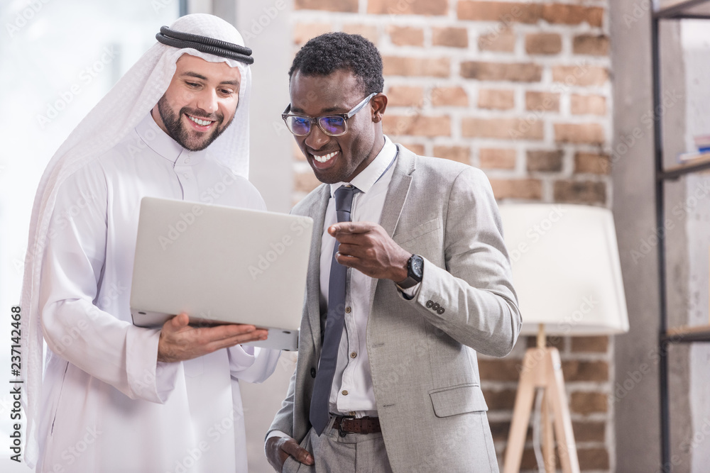 Wall mural african american businessman poiniting at laptop and smiling near arabian partner