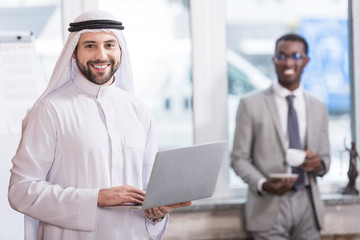 Arabian businessman holding laptop in office with african american partner on background