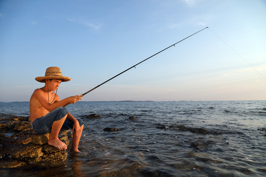 Boy Sitting On The Sea Rocks With A Fishing Rod And Patiently Waiting To Catch A Fish