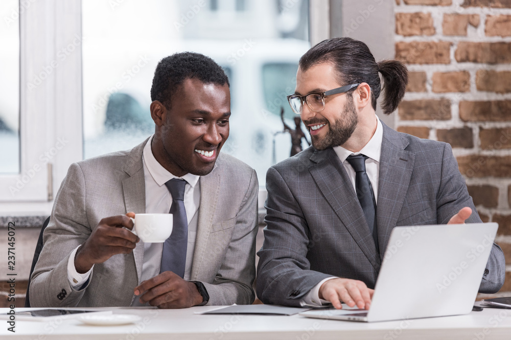 Wall mural smiling multicultural businessmen drinking coffee and looking at laptop in office