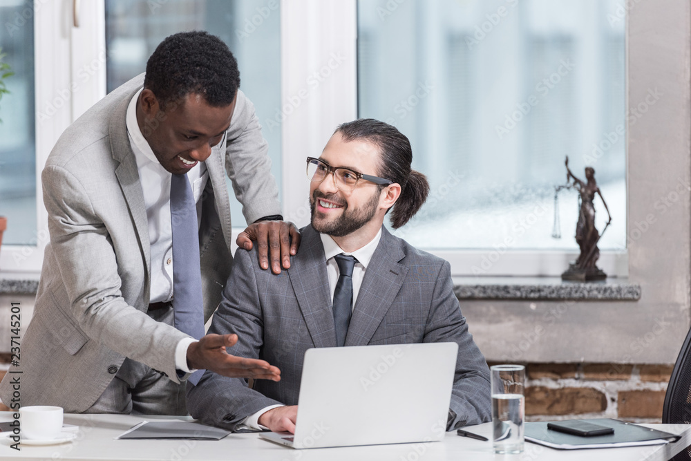 Wall mural smiling african american businessman putting hand on partners shoulder and pointing at laptop in off