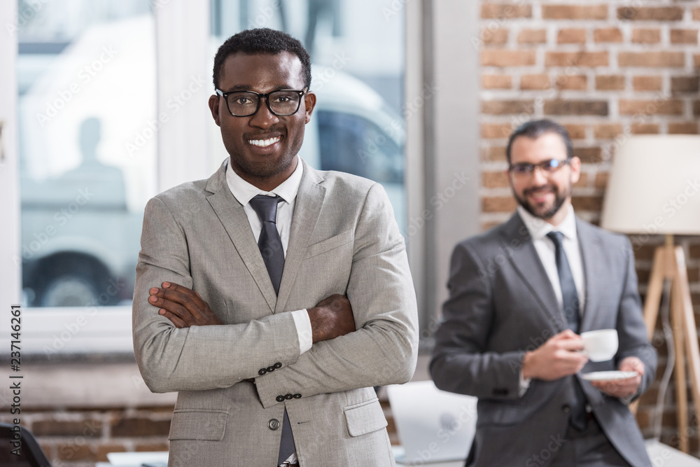 Wall mural smiling african american businessman with arms crossed looking at camera and handsome partner at bac