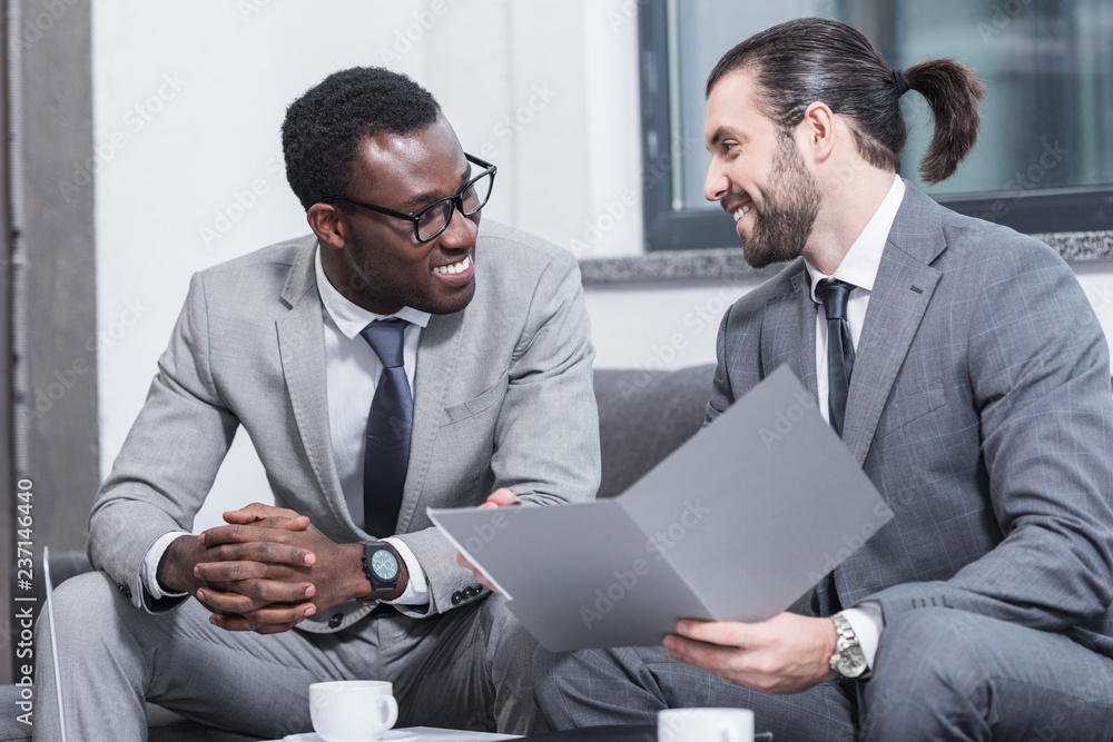 Wall mural smiling multiethnic business partners sitting on couch and talking in office