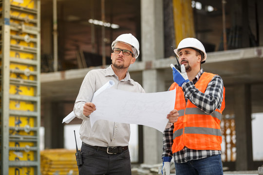 Male architect and developer with walkie talkie and blueprints at construction site