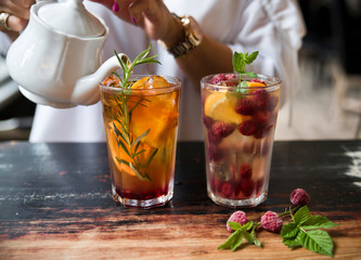 White tea with syrup, orange, raspberry, rosemary and mint on a rustic background. In the photo the hands of the girl are seen, which pours tea from a white vintage teapot.