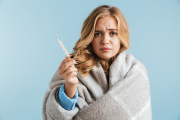 Image of ill upset woman 20s wrapped in blanket holding thermometer, isolated over blue background