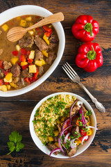 Beef and vegetables stew. White casserole and bowl with meat, bulgur, red onion and fresh parsley on wooden table, top view