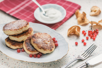 On a white table made of artificial stone is a plate with five cottage cheese pancakes and red berries . On the table is wooden cutting board and a red checkered napkin. Fork and knife.