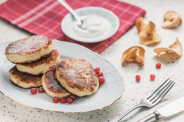 On a white table made of artificial stone is a plate with five cottage cheese pancakes and red berries . On the table is wooden cutting board and a red checkered napkin. Fork and knife.