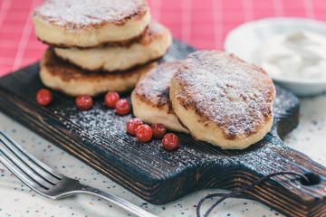 On a white table made of artificial stone is a wooden cutting board with five cottage cheese pancakes and red berries . On the table a red checkered napkin, sour cream, fork and knife.