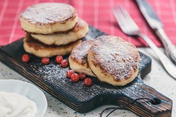 On a white table made of artificial stone is a wooden cutting board with five cottage cheese pancakes and red berries . On the table a red checkered napkin, sour cream, fork and knife.
