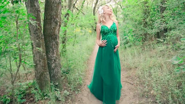 pretty pregnant girl with blond curly hair, decorated with white flowers, walks through the forest alone in a luxurious expensive green dress, gently hugging her tummy slowly walks towards the camera