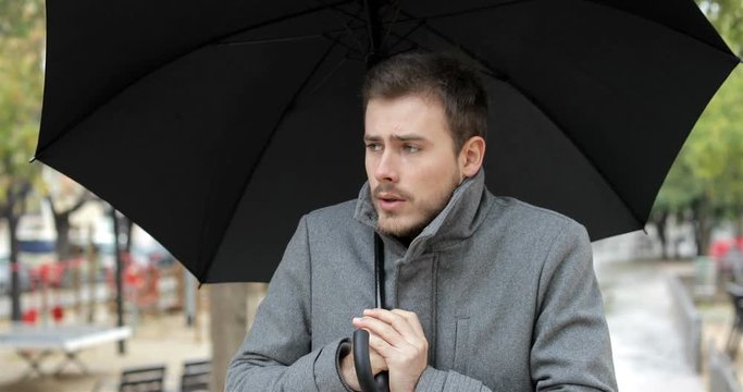 Front view portrait of an angry man getting cold walking towards camera under an umbrella in a rainy winter day