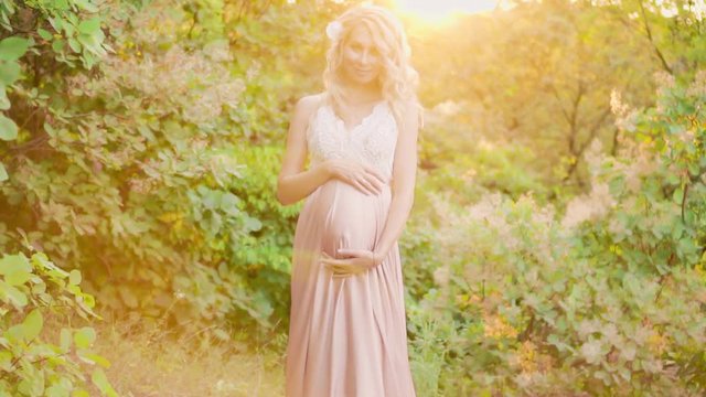 smiling pregnant girl standing in the summer forest and in the rays of the morning sun, dressed in a delicate, superb pink dress in a white lace top, slowly strides forward, embracing her tummy