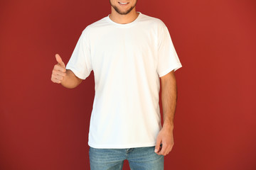 Young man in stylish t-shirt showing thumb-up gesture on color background