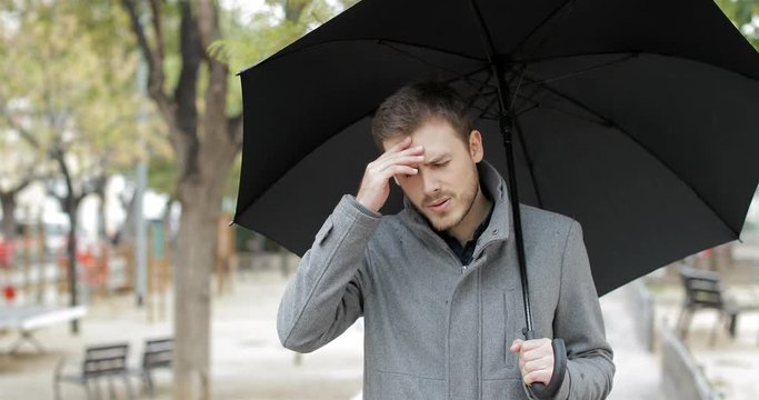 Angry man suffering head ache walking towards camera holding an umbrella under the rain in a park