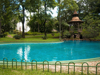 Blue pool in garden with gazebo and trees.