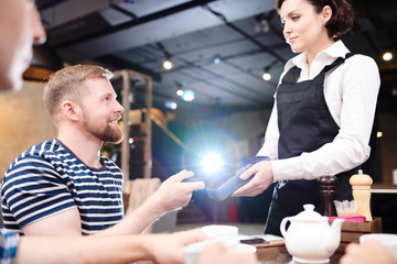 Positive satisfied pub quest in stripped tshirt sitting at table and looking at pretty waitress while paying for lunch with gadget