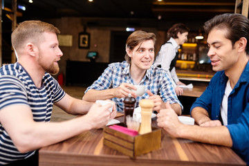 Positive excited young men sitting at table and drinking tea while sharing news at lunch