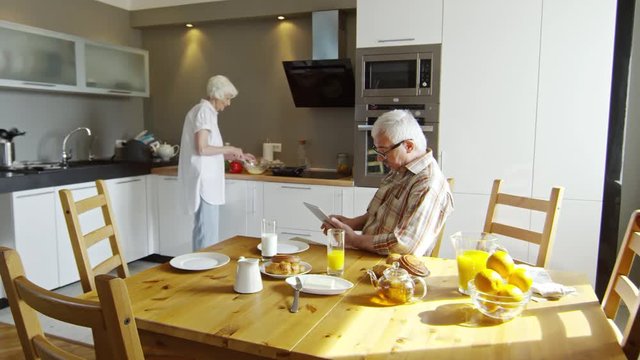 Zoom in of old man using tablet and talking to old woman cooking breakfast in the kitchen