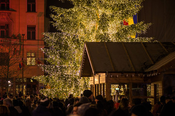 Crowded Christmas market in city center. Decorated Christmas tree with glowing lights and wooden huts full with gifts. Beautiful Christmas background. 