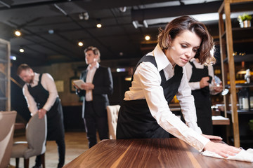 Serious busy young waitress and her colleagues preparing restaurant for open and making cleanup: she wiping wooden table with cloth