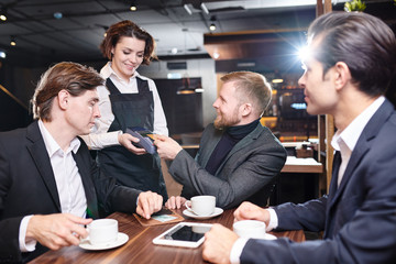 Positive attractive young waitress in black apron standing at table of guests and giving payment terminal to businessman who paying with contactless card in cafe