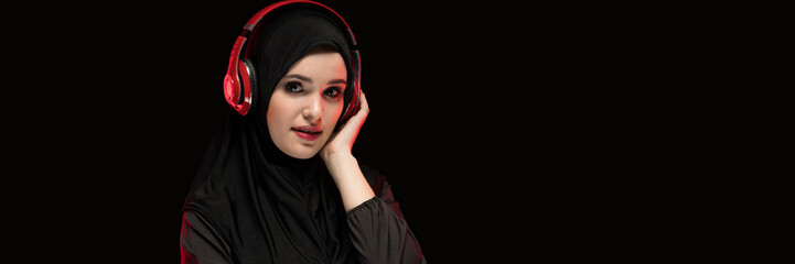 Portrait of beautiful smart young muslim woman wearing black hijab listening to music in headphones with copyspace on black background