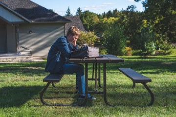 Fototapeta premium Upset teenager sitting at a picnic table in a park all alone. His backpack is lying on the table next to him.