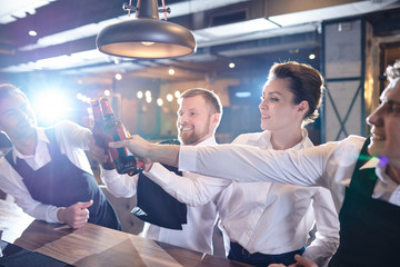 Group of cheerful excited waiting staff in uniform standing at bar counter and clinking beer...