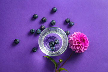 Glass of fresh water with blueberries and flower on color table