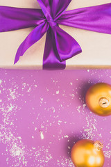 Gift with atlas ribbon and bow-knot, Christmas balls, snow on a violet background. Copy space.