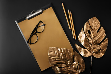 Golden tropical leaves, clipboard and glasses on dark background