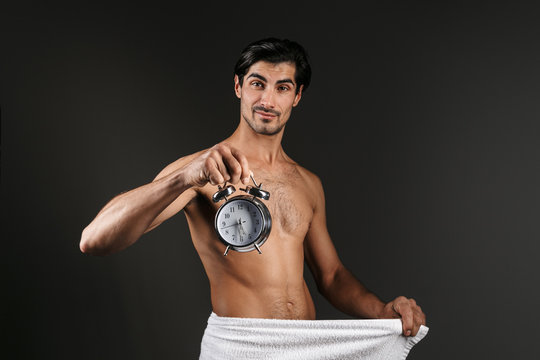 Man isolated over dark background covering his genitals with towel holding alarm clock with half of the sixth time.