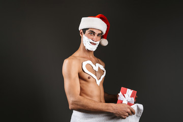 Young santa naked man posing isolated over dark wall background with shaving gel on beard and heart...