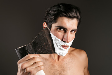 Handsome young naked man posing isolated over dark wall background with shaving gel on beard...