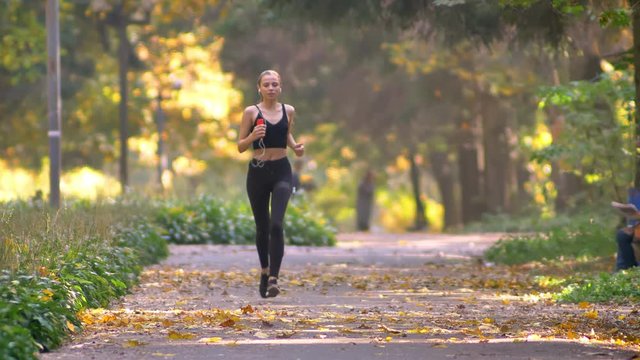 Young girl in jumpsuit with earphones running in autumnal park, stops to look into smartphone and continues jogging.