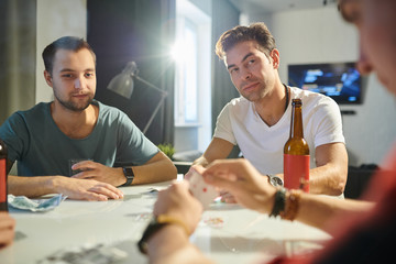 Young men sitting at the table drinking beer and playing cards at home party