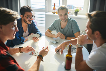 Young men drinking beer at the table at home and playing cards for money