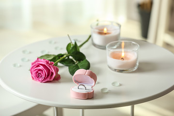 Engagement ring in box with rose and burning candles on white table