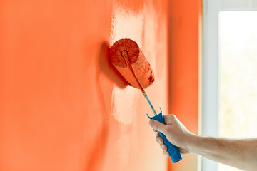 Male hand painting wall with paint roller. Painting apartment, renovating with red orange color...
