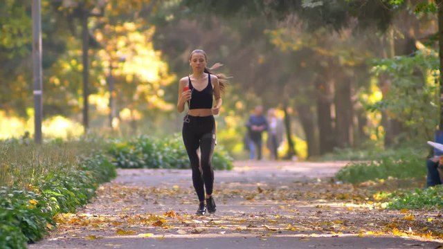 Young girl in jumpsuit with earphones running in autumnal park, stops to tie her snickers and continues jogging.