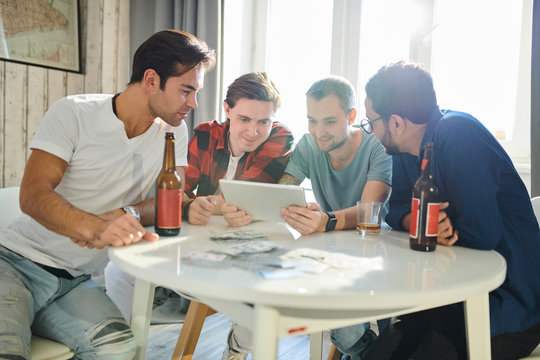 Young men sitting at the table drinking beer and communicating online with their friend or making online order of food using digital tablet