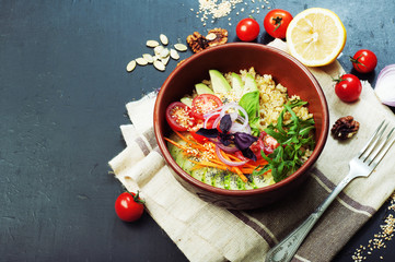 Bowl with bulgur, cherry tomatoes, slices of avocado, cucumber, carrot, arugula and basil on a dark blue surface. Useful and tasty diet food.