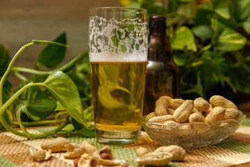 Beer in bottle and glasses and peanuts in crystal bowl. Table with vintage bamboo mat and liana vine in the background.