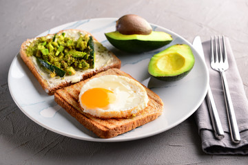 Plate with toasted bread, avocado and fried egg on grey table