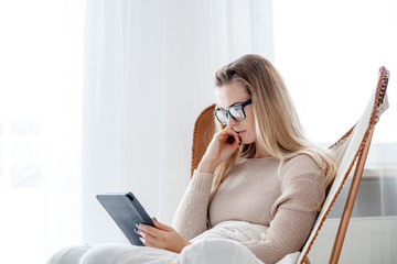 Young woman in glasses reading ebook using digital reader while sitting in comfortable armchair at...