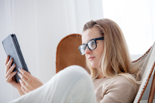 Young woman in glasses reading ebook using digital reader while sitting in comfortable armchair at home
