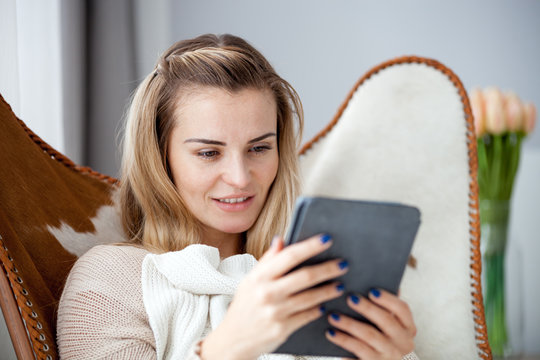 Young woman reading ebook using digital reader while sitting in comfortable armchair at home
