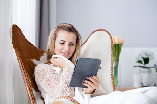 Young woman reading ebook using digital reader while sitting in comfortable armchair at home